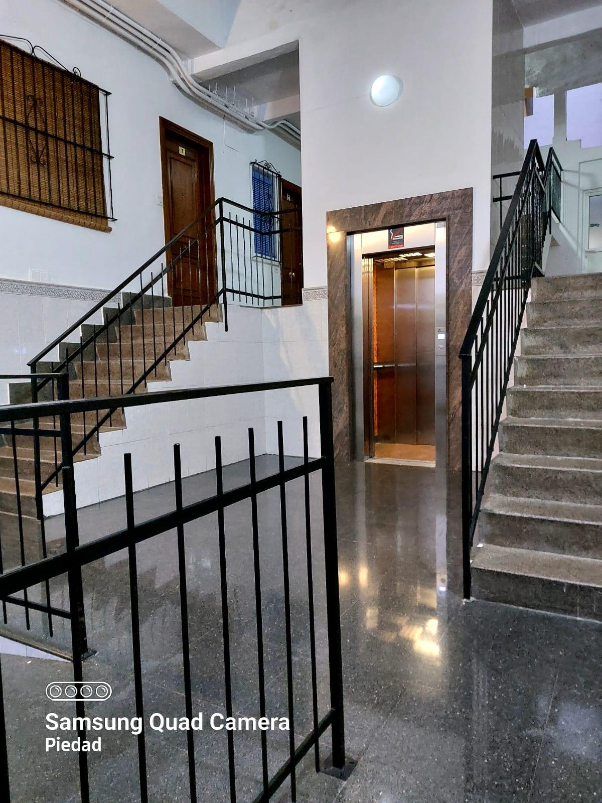 Interior building lobby showing two staircases on either side and a central elevator with a marble-framed doorway; polished floor and white walls.
