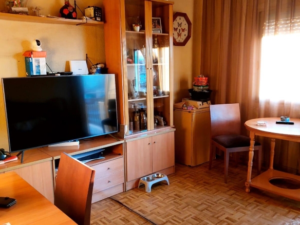 Flat-screen TV on a wooden cabinet with a glass-front display cabinet nearby, round dining table, and brown curtains in a warm living room.