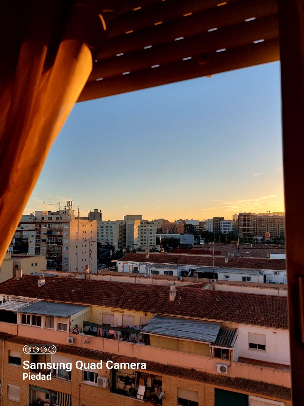 Sunset over a city skyline viewed from a window, with a brown curtain on the left side.