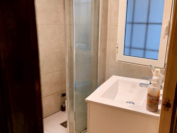 Small bathroom with a glass sliding shower door, white sink, and towel hung above the shower. A frosted window is to the right with toiletries on the sink.
