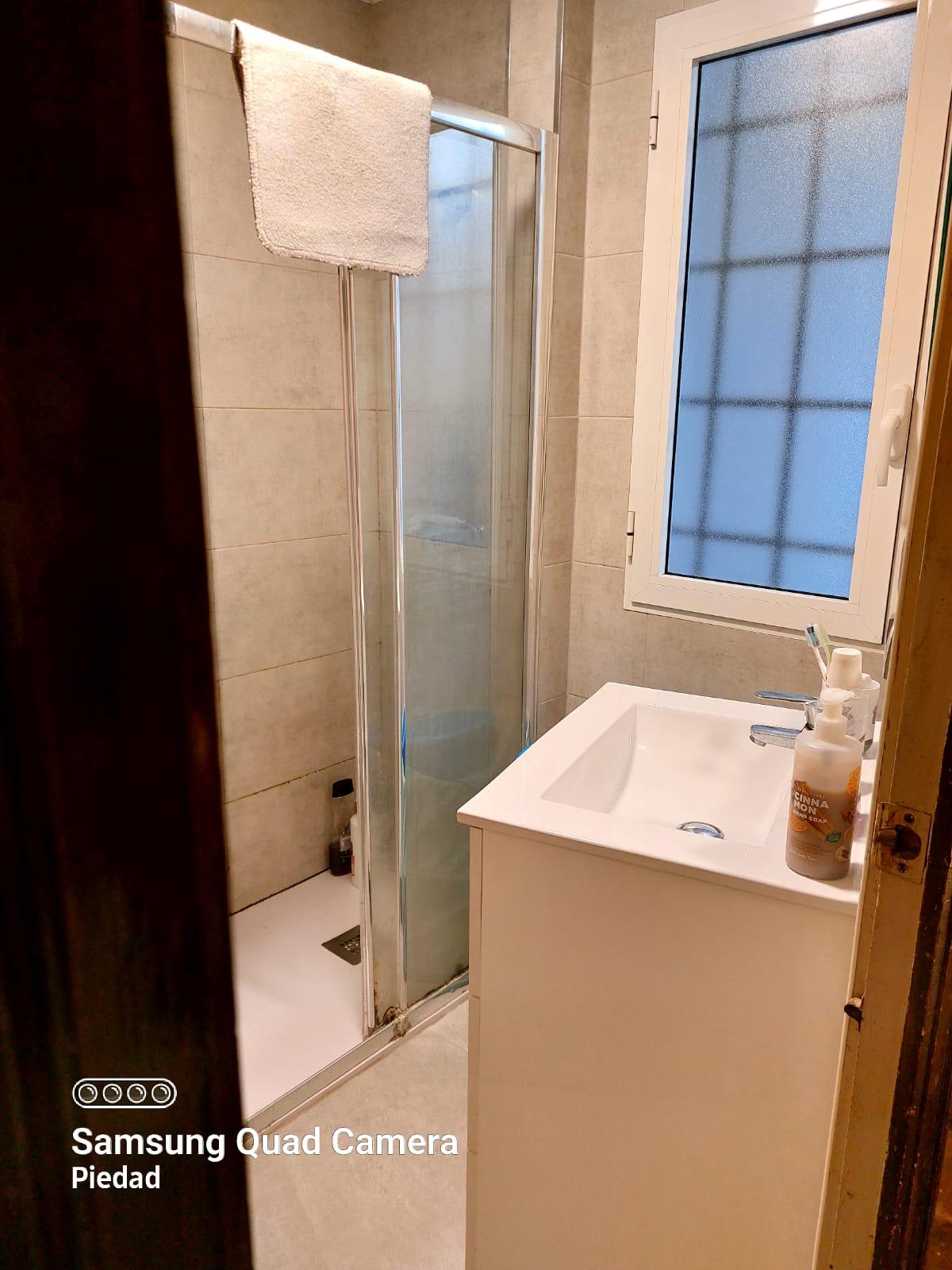 Small bathroom with a glass sliding shower door, white sink, and towel hung above the shower. A frosted window is to the right with toiletries on the sink.
